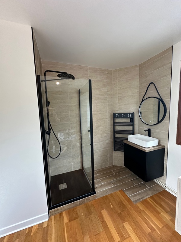 salle_de_bains Salle de bain rénovée à Osny (Val-d’Oise), au style contemporain et épuré. Revêtement mural effet béton, meuble en bois clair avec vasque moderne et miroir rétroéclairé. Baignoire îlot design, niche murale lumineuse et touche végétale suspendue pour une ambiance zen.